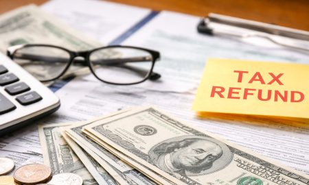 Primer plano de billetes y monedas de dólares estadounidenses, una calculadora y documentos fiscales del IRS con una nota de “Tax Refund” sobre el papeleo.