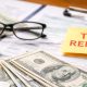 Primer plano de billetes y monedas de dólares estadounidenses, una calculadora y documentos fiscales del IRS con una nota de “Tax Refund” sobre el papeleo.